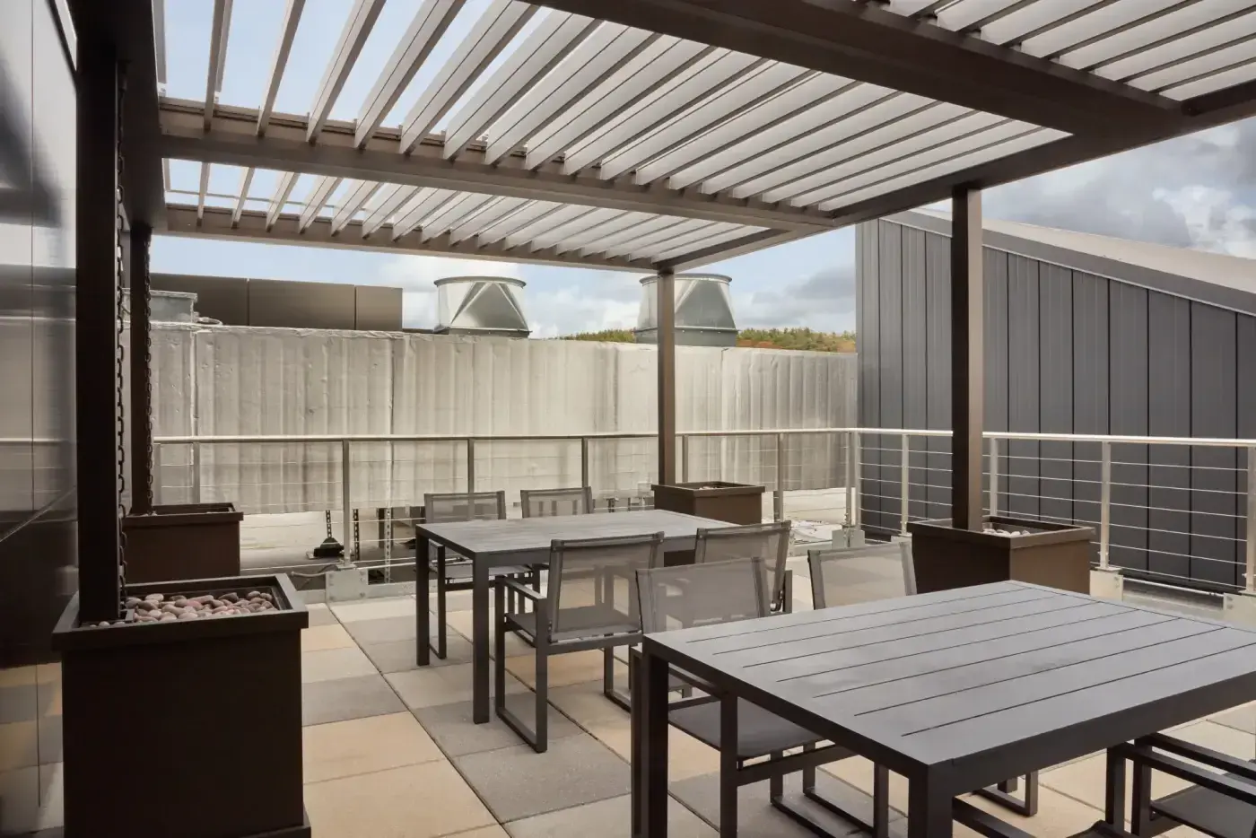 This stunning roof deck employee gathering area is 12' 6" wide by 20' long. The Apollo opening roof's unique system has remote control easy opening and closing operation, providing variable shading and light, and protection from inclement weather. Shade louvers feature a clear coat finish, while the posts, frame, and gutters have an anodized bronze finish. The securing planter box anchors, crafted in solid cellular vinyl with stainless steel liners, are 24" square by 30" H. Walpole's custom color matches the anodized bronze frame.