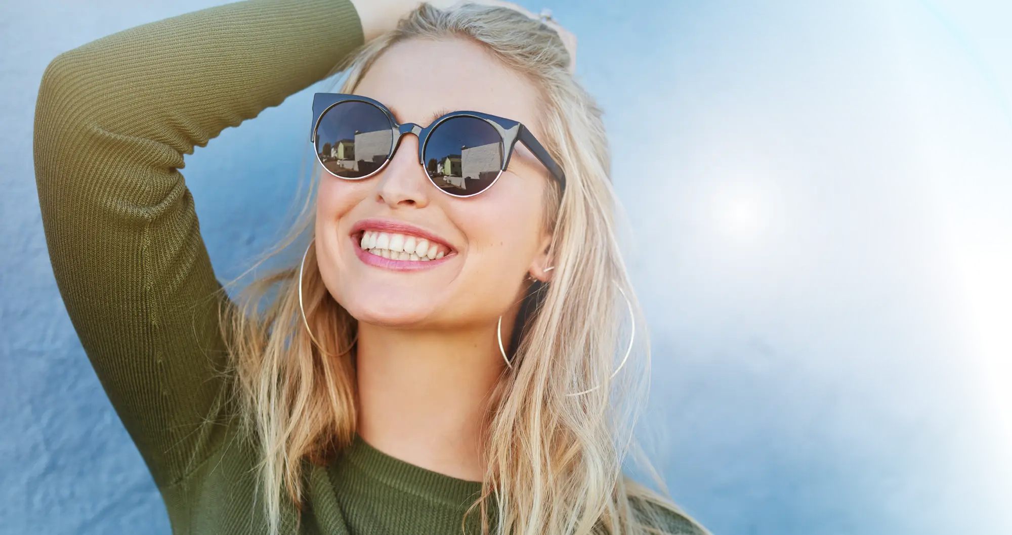 Woman standing against a wall, hand in her hair, smiling with sunglasses on.