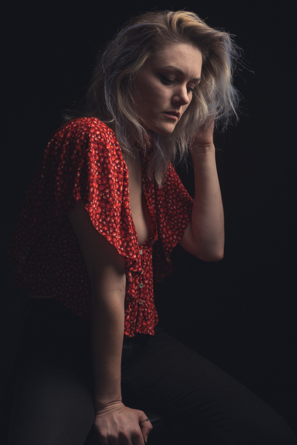 a portrait of Ferring wearing a red blouse, she is sitting on a stool with her hand leaning on the stool and one arm in her hair. she is sitting against a black background.