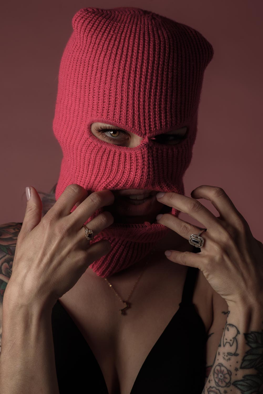 a portrait of Christina on a pink background, she is wearing a black bra and a pink ski mask and she is stretching out the mask mouth opening.