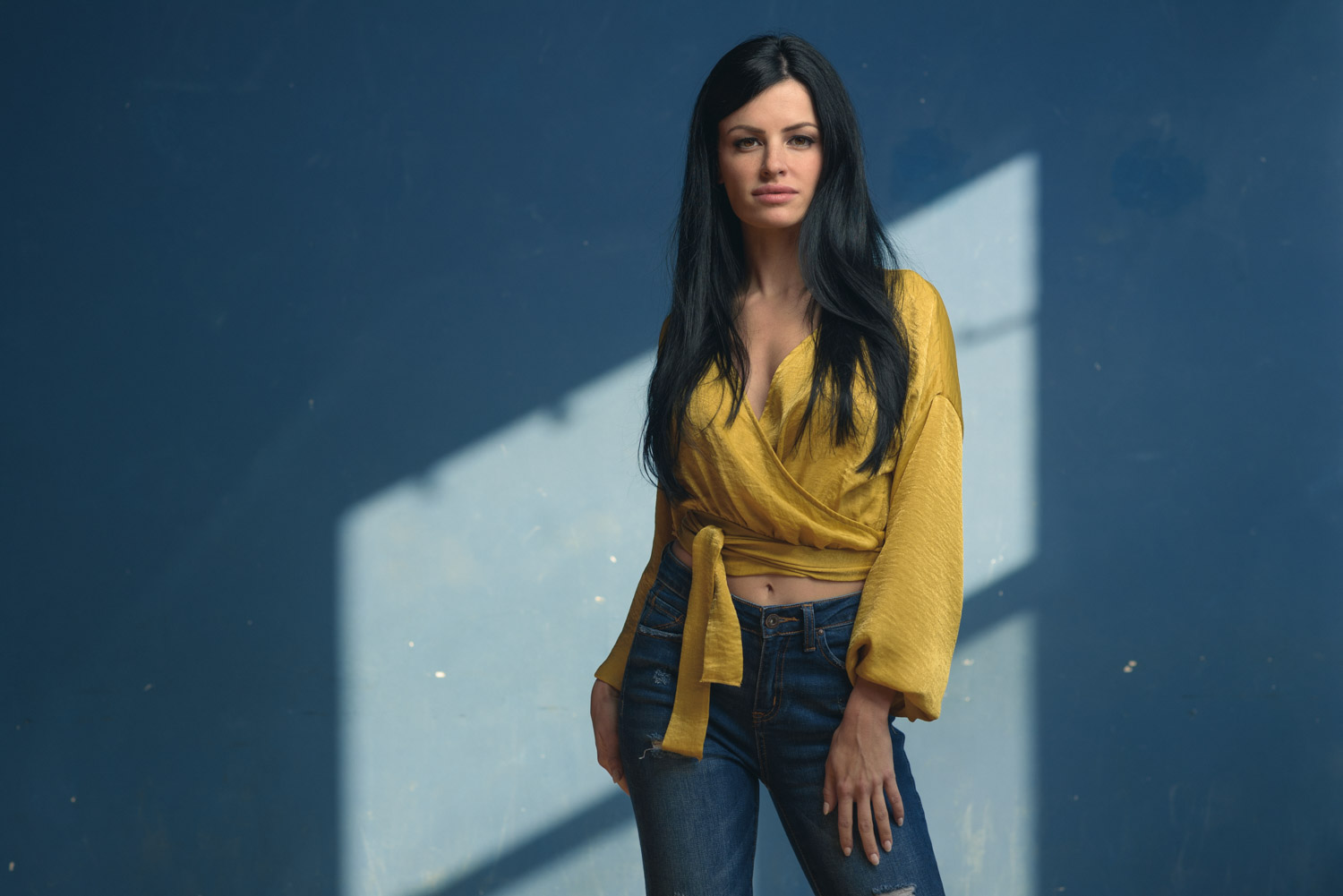 a portrait of Katy wearing a yellow blouse and blue jeans, she is posing in front of a blue wall and the sun is casting a reflection of the window upon the wall behind her.