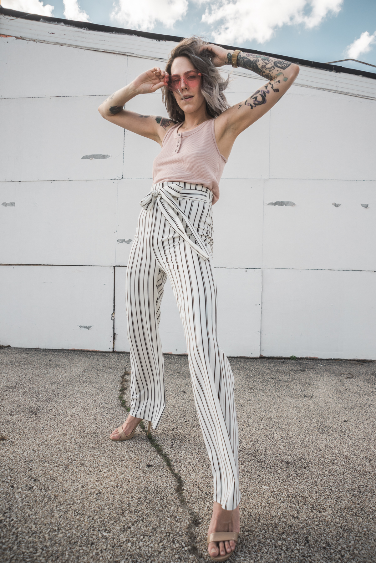 portrait of Christina in pin striped pants in front of white wall with blue sky peaking right above the wall and shot from a low angle