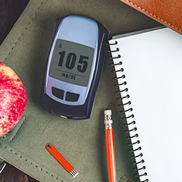 Glucometer reading 105 placed on the table along with an apple, pencil and note.