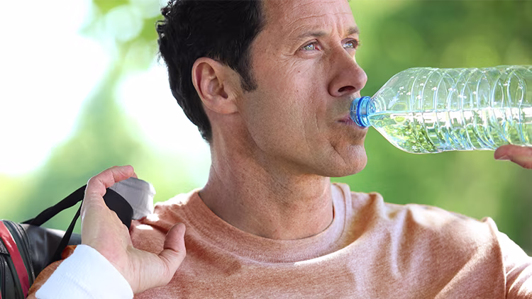 A man drinking water carrying a bag post workout and shades of trees in background.