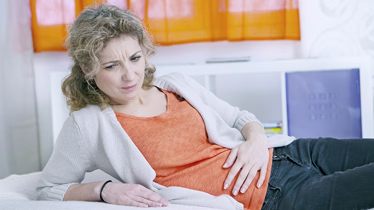 A woman in orange t-shirt laying on bed holding her stomach in one hand in pain.