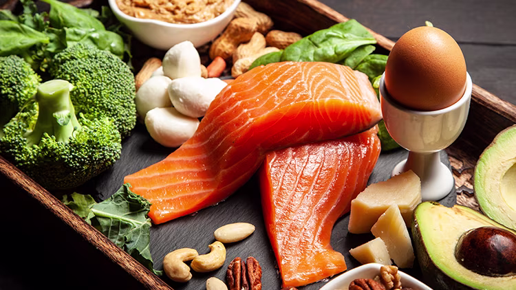 Two pieces of salmon placed at centre surrounded by an egg, broccoli, sliced avocado, mushroom, cheese pieces and mix of nuts arranged on wooden tray.