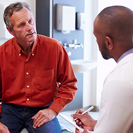 A man in orange shirt talking to doctor while Doctor taking notes.