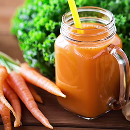 A jar of carrot juice placed in between cut pieces of lemon, ginger, a bunch of carrots and lettuce on a wooden table.