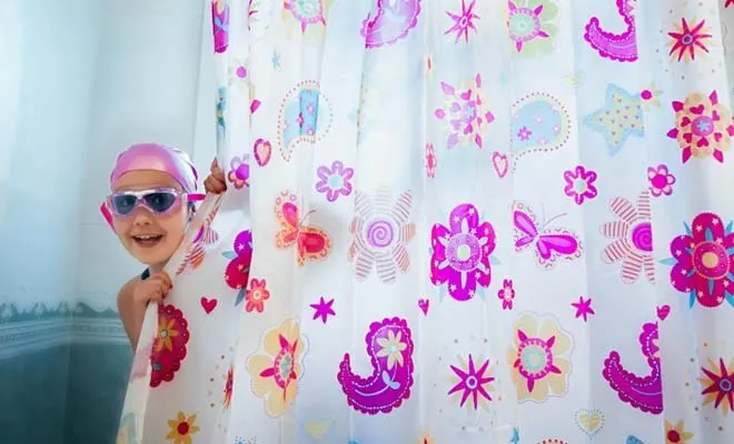 Une fillette portant un bonnet de douche et des lunettes de natation se penche la tête hors d'un rideau de douche imprimé de fleurs mauves.