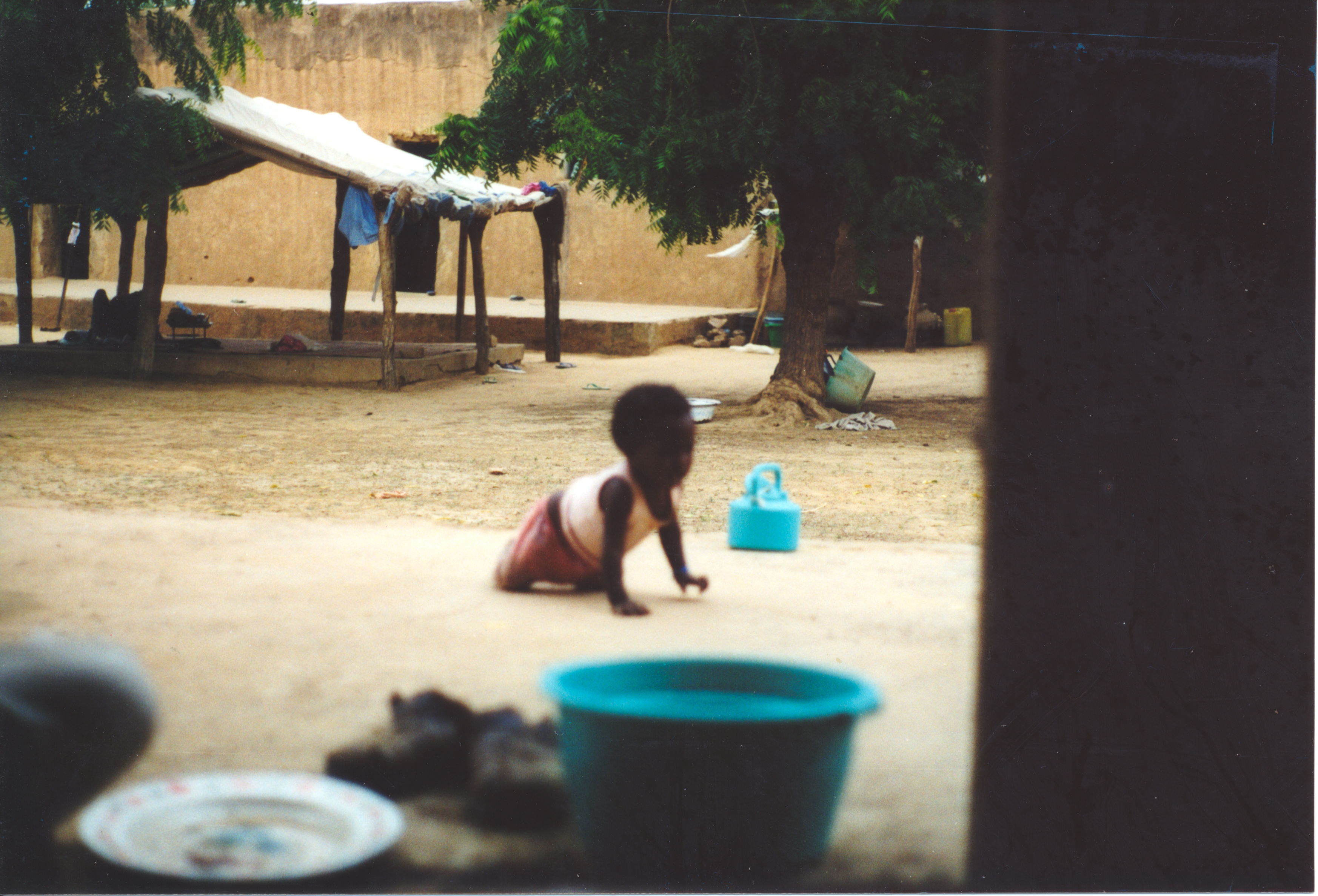 A baby crawls on a cement platform in family compound. A mud-brick building is in the background. In front of that are two trees and a pavilion for shade.