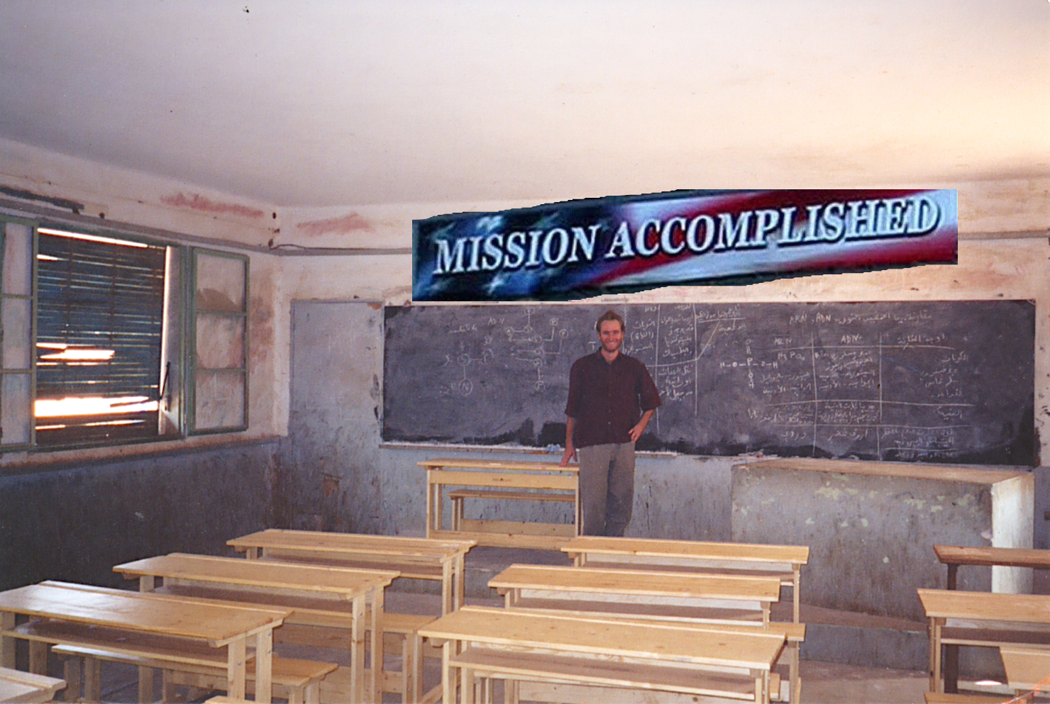 Altered photo. 'Mission Accomplished' banner from President Bush's aircraft carrier speech superimposed on original photo. John is standing at the head of a classroom facing the viewer. Behind him is a dusty chalkboard nearly the length of the wall. In front of him stand several bench-style desks, for two kids each. The walls are dirty; paint is chipping off surfaces; the shutters in the window are blocking the sunlight except where they are broken.