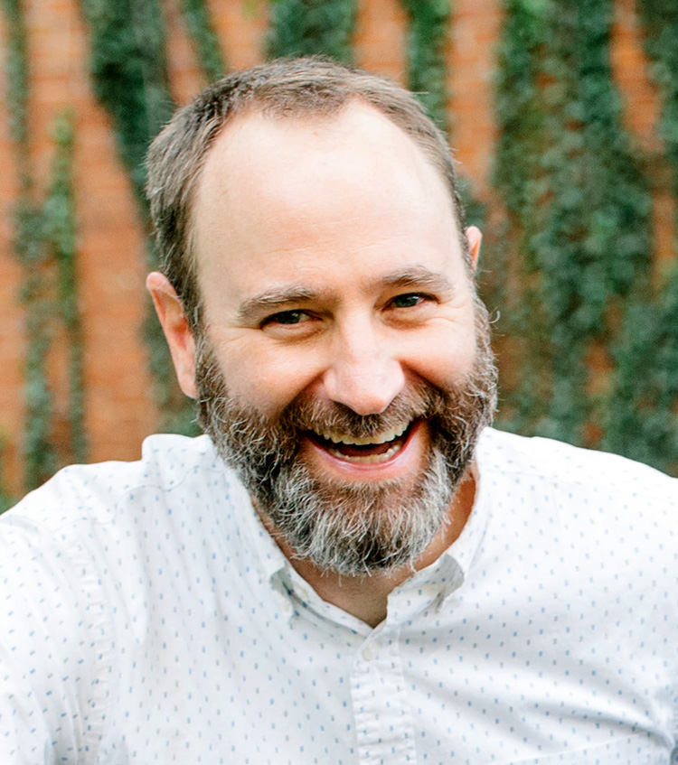 John Osterman. Physical description: a White man with short brown hair and a graying beard, smiling outside in front of a brick wall with ivy on it.