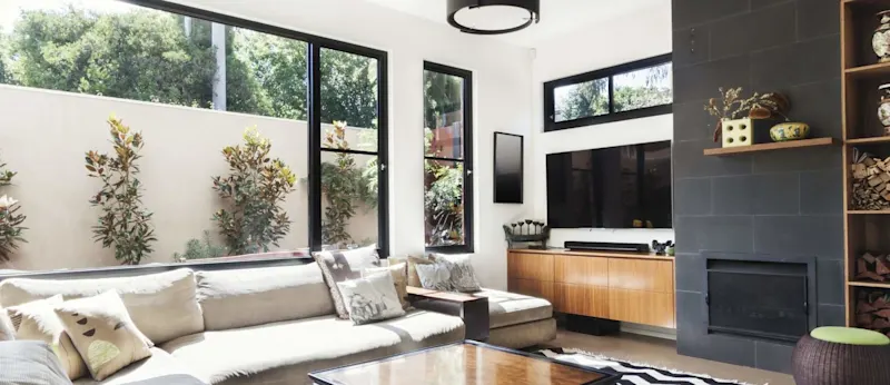 A modern, well-furnished living room with large windows, a stylish fireplace, and lush greenery outside.