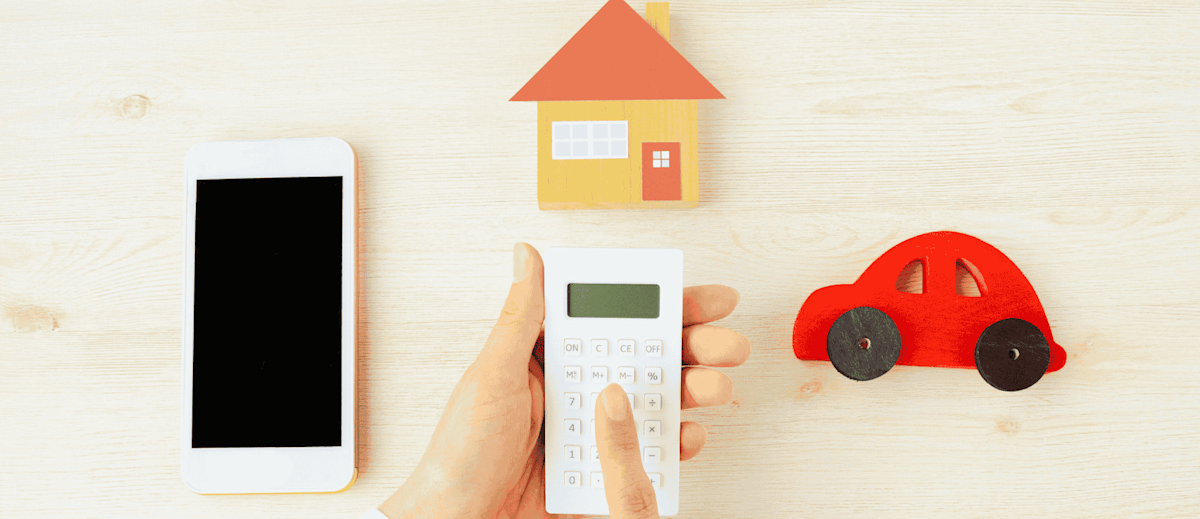 A person using a calculator next to a phone, model home, and a model car.