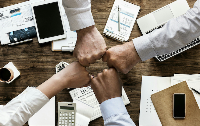 Four people engaging in a fist bump overtop of insurance and financial documents on a crowded desk.