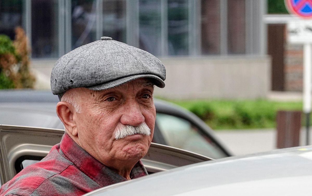 An elderly driver wearing a hat and red checkered shirt getting into a vehicle.