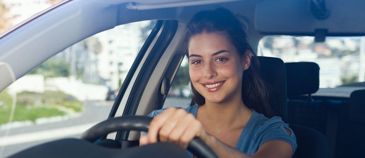 A smiling young woman sitting in the driver's seat of a car with her hands on the steering wheel.