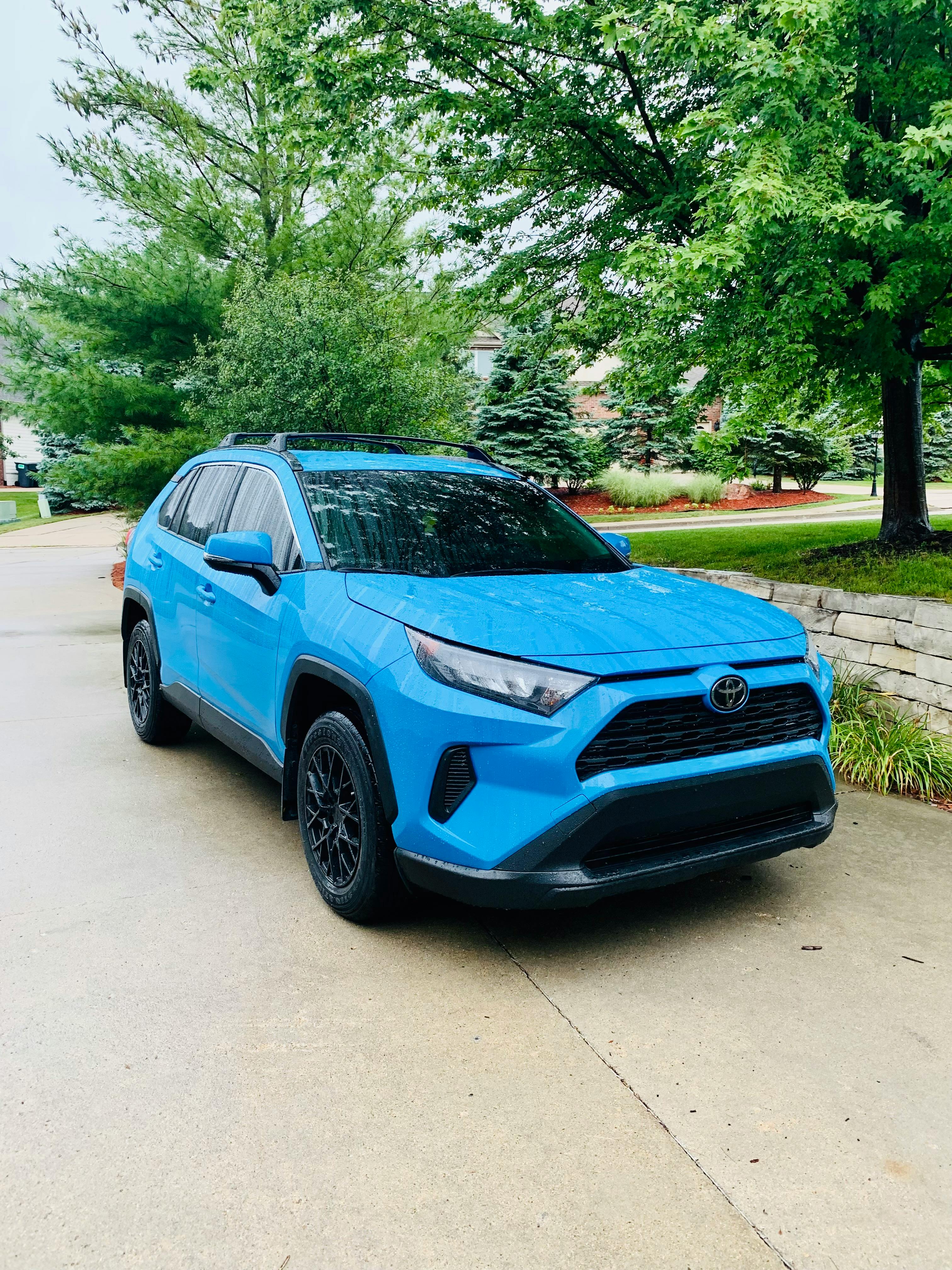 Front view of a bright "Blue Flame" Toyota RAV4 Adventure SUV with black wheels and roof racks, parked on a suburban driveway.
