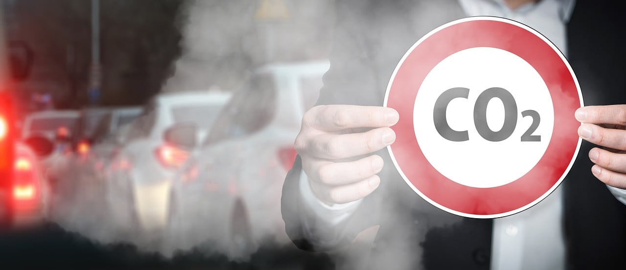 A man holding a CO2 sign in a line of traffic with car exhaust blowing over him.