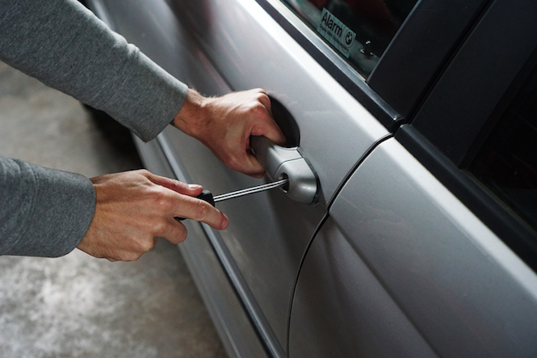 A close-up of a man breaking into a car with a screwdriver, representing the risk of theft and break-ins at Auckland Airport car parks that Kiwi travellers need their car insurance to cover.