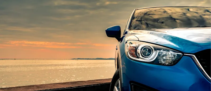 A front-angle view of a blue car parked near the ocean at sunset, capturing the total experience of car ownership and running costs in Auckland for 2026, as illustrated by Quashed.co.nz.
