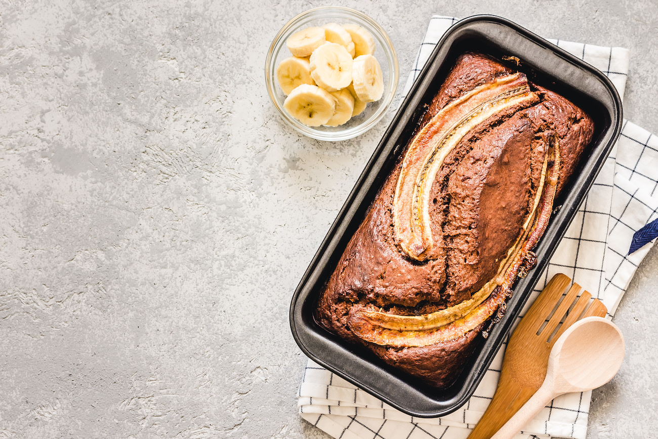 Banana bread in tin with bananas and wooden utensils