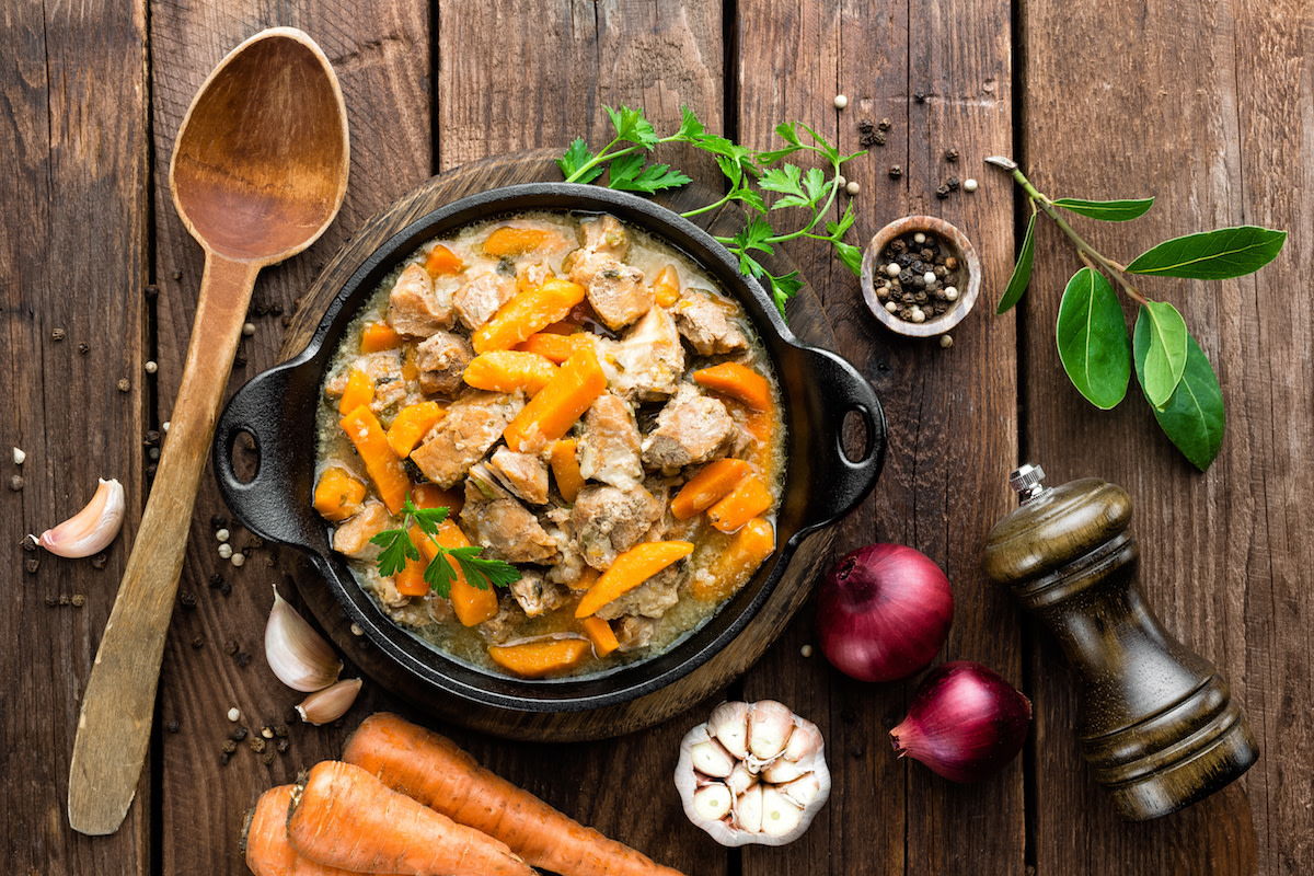 Stew of meat and carrots in pot with raw ingredients on wood