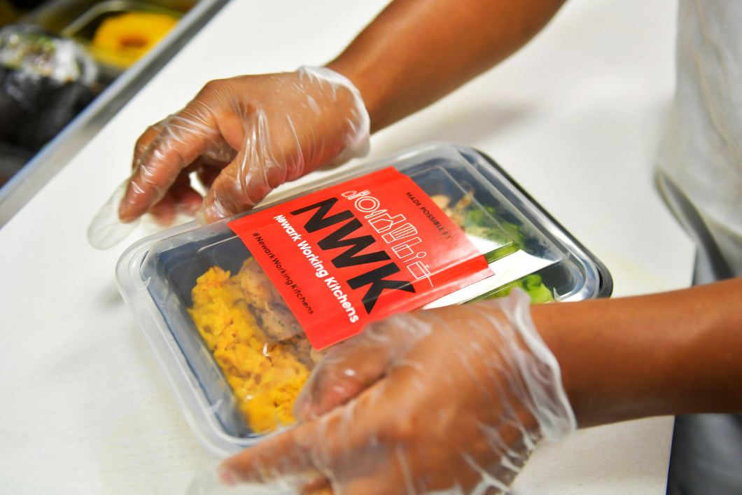 A close up look at gloved hands sealing a package that contains a Newark Working Kitchen meal. The NWK logo is prominent on the meal package's cover.