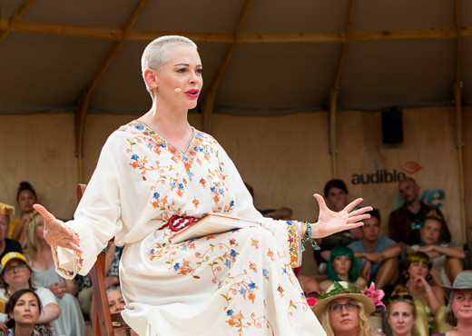 Rose McGowan sits on a chair on the Audible Live Stage at the Wilderness Festival. Her arms are spread wide, her fingers splayed as she performs. She is wearing a white dress with embroidered flowers.  