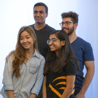Four college interns stand in a group posing for a photo against a neutral background. They are all smiling and happy. One of them is wearing a t-shirt with the orange Audible chevron on it. 