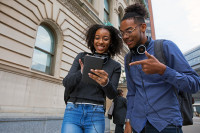 Two high school students stand on a Newark street, earphones around their neck, looking excitedly at a tablet one of them is holding.