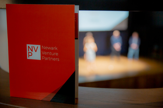 An orange pamphlet with NVP logo + "Newark Venture Partners" in white text is standing on a ledge, while you see figures of people standing on stage in the background.