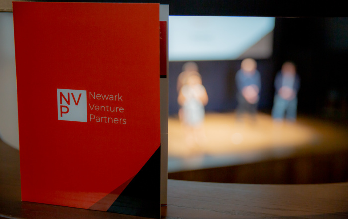 An orange pamphlet with NVP logo + "Newark Venture Partners" in white text is standing on a ledge, while you see figures of people standing on stage in the background.