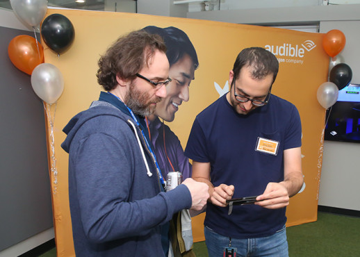 Two men stand close looking down at one of the men's phones. They are in front of a large orange poster displaying a man listening to Audible who appears, due to the angle of his head and that it falls between the two mens' shoulders, to be peering at their phone too.