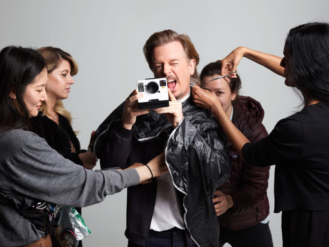 David Spade stands center with a polaroid camera in hand, snapping a photo, while four female stylists surround him prepping his hair and clothing for a photo shoot.