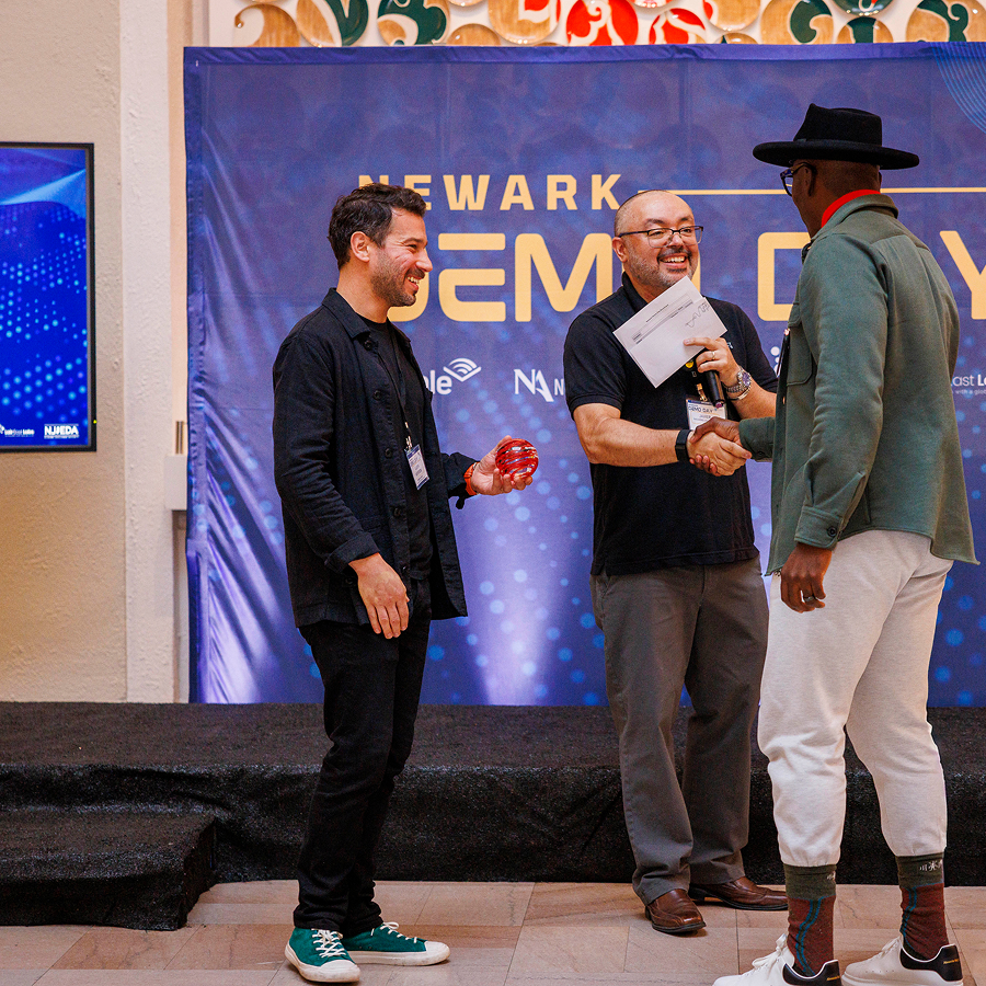 Two men accept a congratulations from another man in front of a Newark Demo Day backdrop. 