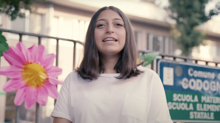A woman stands in front of a school in Italy talking to camera (this is a screenshot from a video). There is a fence behind her and on it is a large paper flower with pink petals and a yellow middle as well as a wide blue sign with the school's information in Italian.