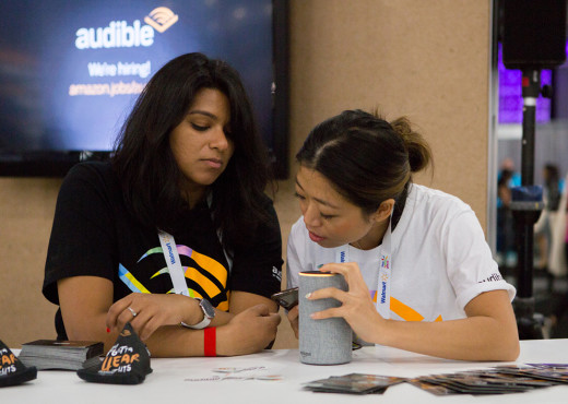 Two women huddle together at a table looking at an Alexa device.