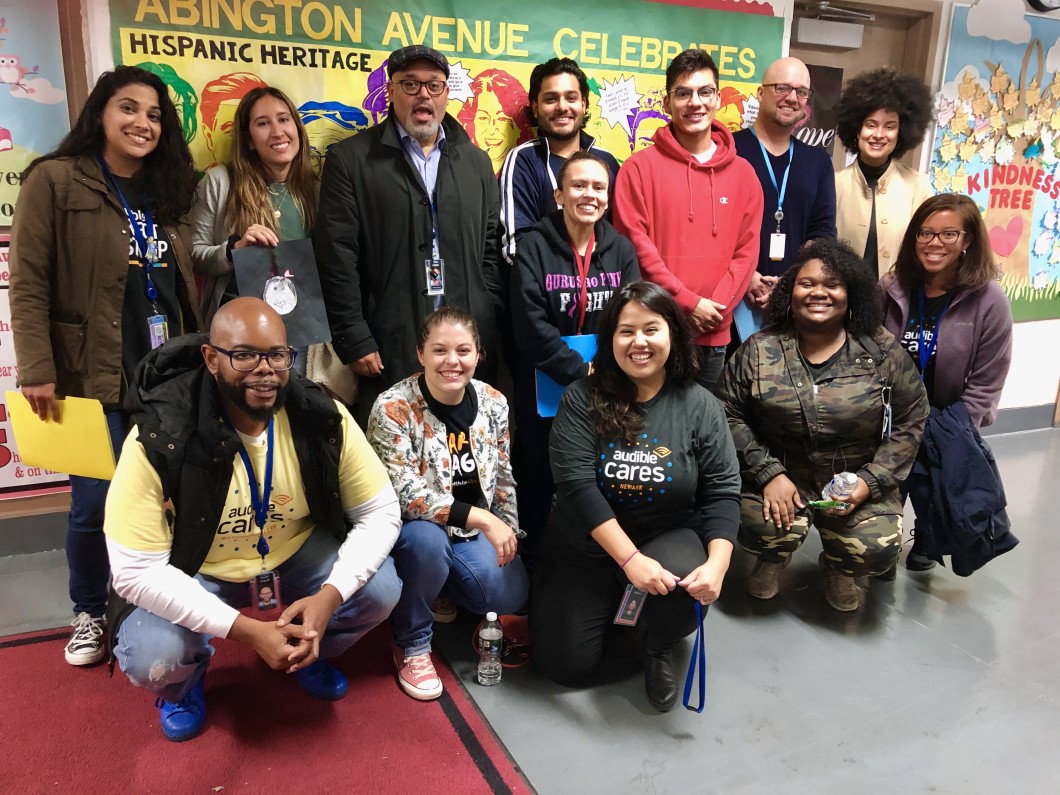 13 Audible employees are standing in front of some student artwork in Abington Avenue Elementary School commemorating notable Latinx figures, the most visible being Sonia Maria Sotomayor, Associate Justice of the Supreme Court of the United States. 