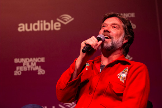 Rufus Wainwright speaks into a microphone at the Sundance event, "A Conversation with Rufus Wainwright." He's wearing a red button down and sitting in front of Audible signage.