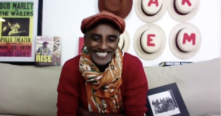Chef Marcus Samuelsson sits on the couch in his department talking on camera for the SXSW panel. He is wearing a maroon sweater and cap and a multicolored scarf. A Bob Marley poster and hats with letters on them that spells out Harlem hang behind him.