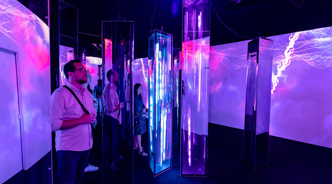 A man with glasses is standing in a dark art installation in which purple light is being reflected on mirrors placed throughout the small room.