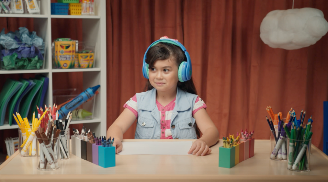 A photo of a young girl wearing large blue headphones. She’s seated at a desk which is covered in a colorful selection of Crayola markers, colored pencils and crayons. 