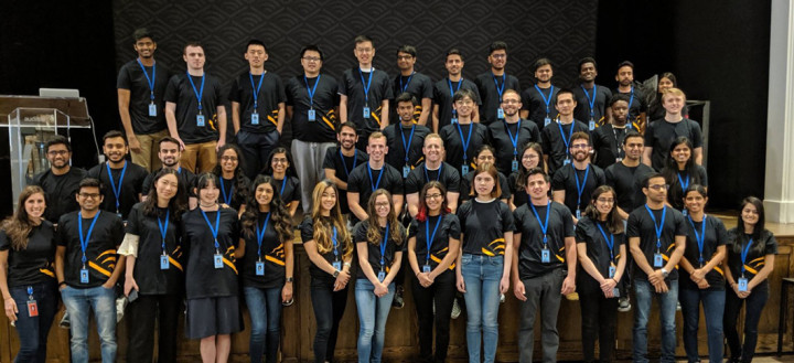 Approximately 30 Audible college tech interns stand lined up four rows deep smiling for a group photo. They all wear black t-shirts with the orange Audible chevron logo on it and wear blue lanyards around their necks with their work badges.