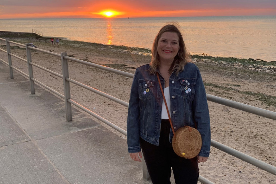 Audible employee Jess Falconer stands on a beach boardwalk. Behind her is a railing and then below it a sandy beach. The sun is setting over the ocean, a red burning glow in the distance. Jess is looking straight to the camera, smiling, while wearing a white t-shirt, jean jacket, black pants and a crossbody straw bag. 