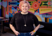 Woman with curly pink hair, smiles at the camera with her hands in her pockets. A colorful wallpaper and tables and chairs are behind her.