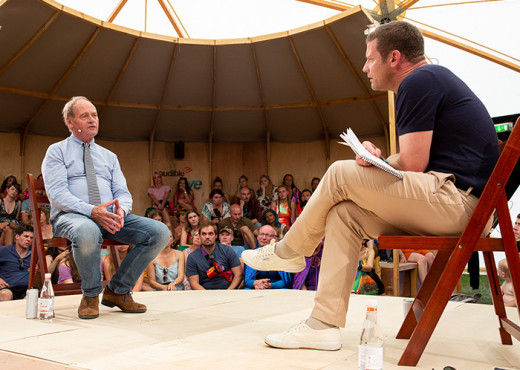 Producer John Lloyd sits in a chair on the Audible stage at the Wilderness Festival in a blue button shirt, tie and blue jeans being interview by Dermot O'Leary who sits on stage in a chair opposite him in a black t-shirt and khaki pants. The audience is viewable in the background.