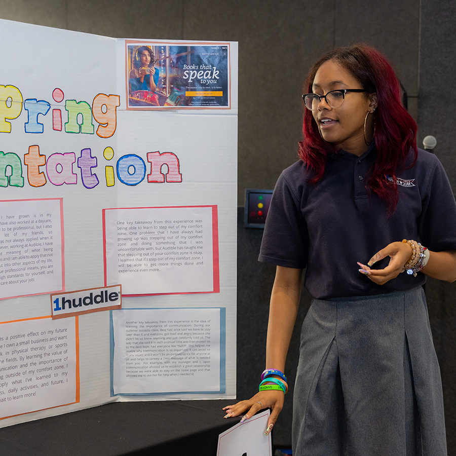 A student stands next to a large piece of posterboard presenting the information pasted on it. 