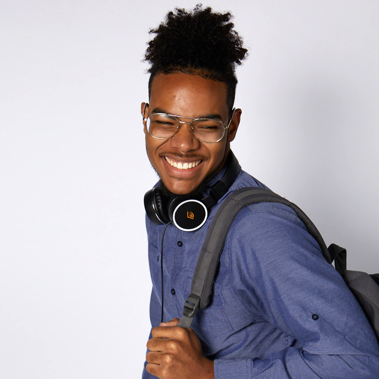 A teen stands sideways against a neutral background facing toward the camera with a big smile on his face. He has a blue button down shirt on, Audible headphones sitting around his next, and a grey backpack over his left shoulder. 