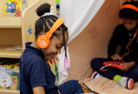 A photo of a young student using an "Audible Story Studio." She's wearing orange headphones with an Audible logo on the side. A second student is using a tablet and headphones in the background.